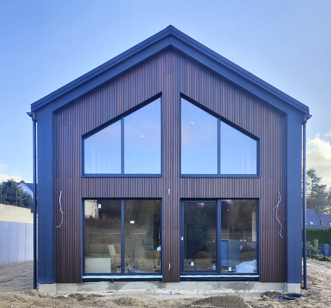 Nowoczesny dom z drewnianą elewacją i dużymi oknami trójkątnymi oraz przesuwnymi drzwiami. Budynek w trakcie wykończenia, widoczne kable i przygotowany teren.