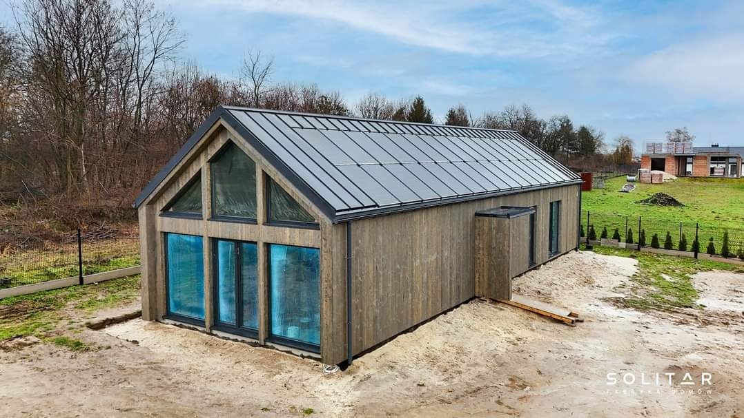 Nowoczesny dom modułowy z drewnianą elewacją i dużymi oknami, widok z zewnątrz w słoneczny dzień. W tle zielony trawnik i inne budynki.