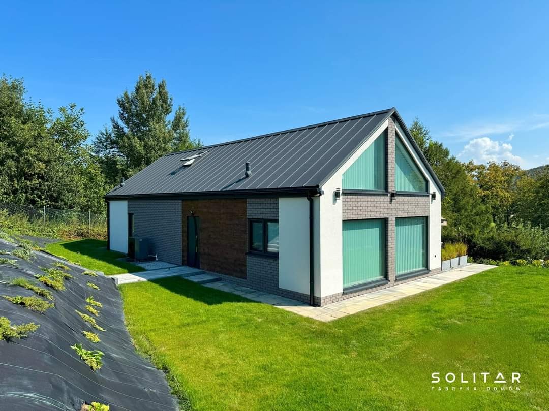 Nowoczesny dom z szarą blachą na dachu, białymi ścianami i elementami z cegły, otoczony zielonym trawnikiem i drzewami w słoneczny dzień.