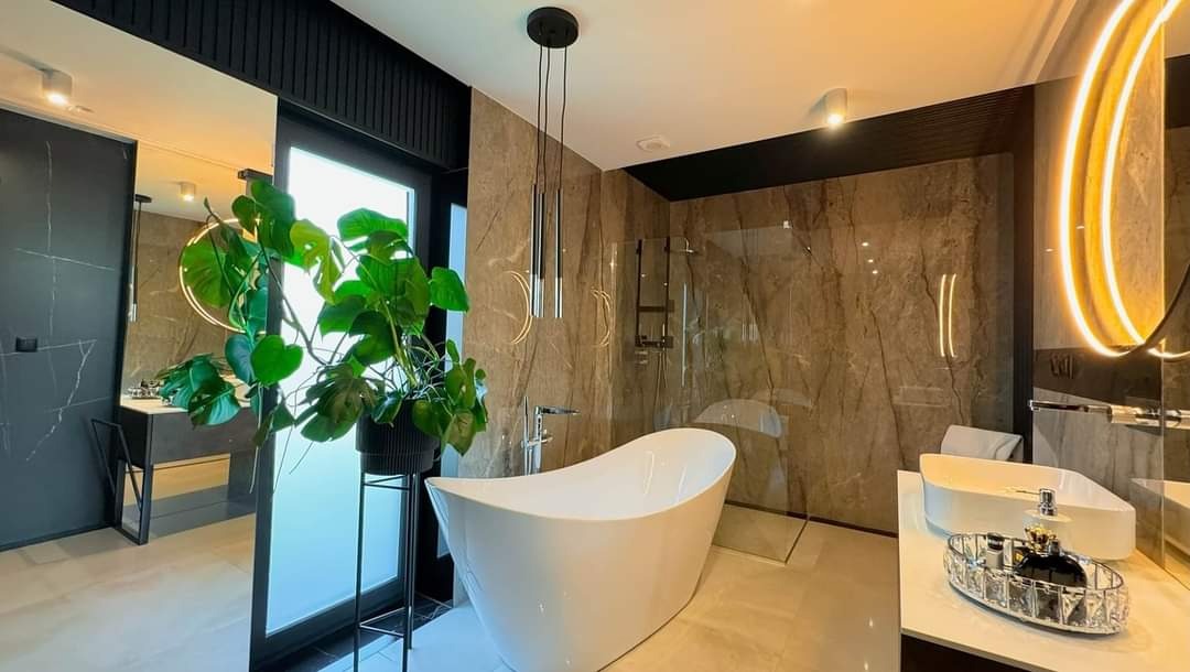 Elegancka łazienka z wanną wolnostojącą, marmurowymi płytkami i nowoczesnym oświetleniem. Roślina doniczkowa dodaje naturalnego akcentu. Widoczne lustro z podświetleniem.
