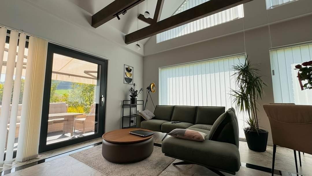 Jasne wnętrze domu z dużym oknem na ogród. Wystrój z drewnianymi belkami, szarym narożnikiem i okrągłym stolikiem kawowym. Nowoczesny design.