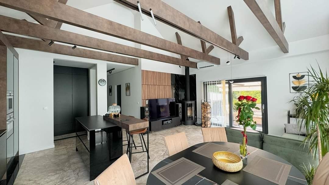 Jasne wnętrze domu z drewnianymi belkami stropowymi, kominkiem i nowoczesnym wyposażeniem. Widoczne krzesła barowe i stół z kwiatami. Nowoczesny design.