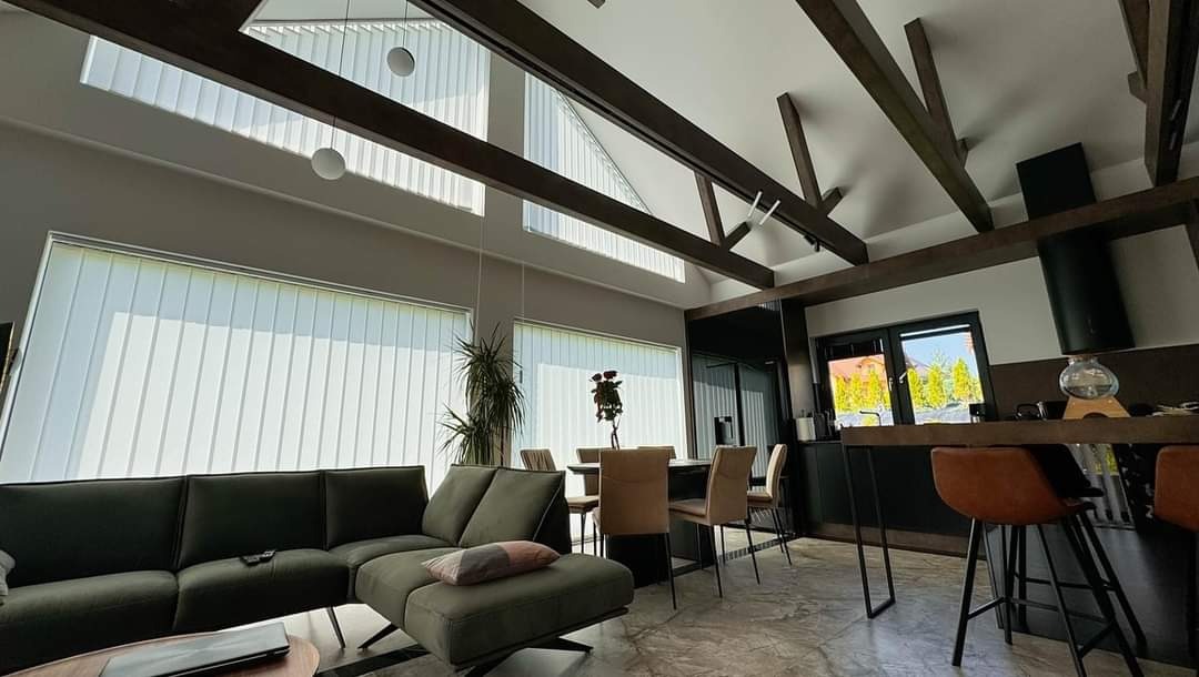 Jasne wnętrze domu z drewnianymi belkami stropowymi i pionowymi żaluzjami w oknach. Nowoczesny salon z kuchnią i jadalnią w szarościach i brązach.