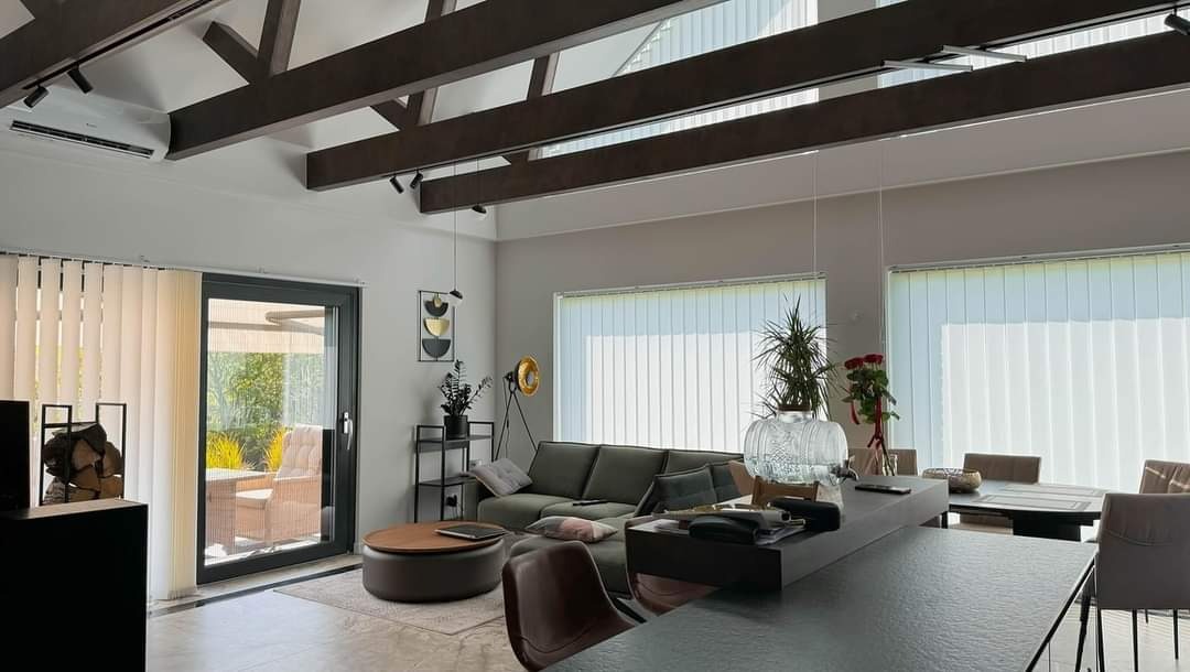 Jasne wnętrze domu z widocznymi drewnianymi belkami stropowymi, szarym narożnikiem i dużymi oknami z pionowymi żaluzjami. Na zewnątrz taras z meblami ogrodowymi.
