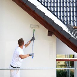 Piotrek donski super ceny dekarz i Cieślak.pl - Mężczyzna maluje białą elewację domu wałkiem na teleskopowym kiju, widoczna część dachu z ciemną dachówką i drewnianą podbitką, rusztowanie w tle.