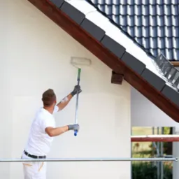 Mężczyzna maluje białą elewację domu wałkiem na teleskopowym kiju, widoczna część dachu z ciemną dachówką i drewnianą podbitką, rusztowanie w tle.