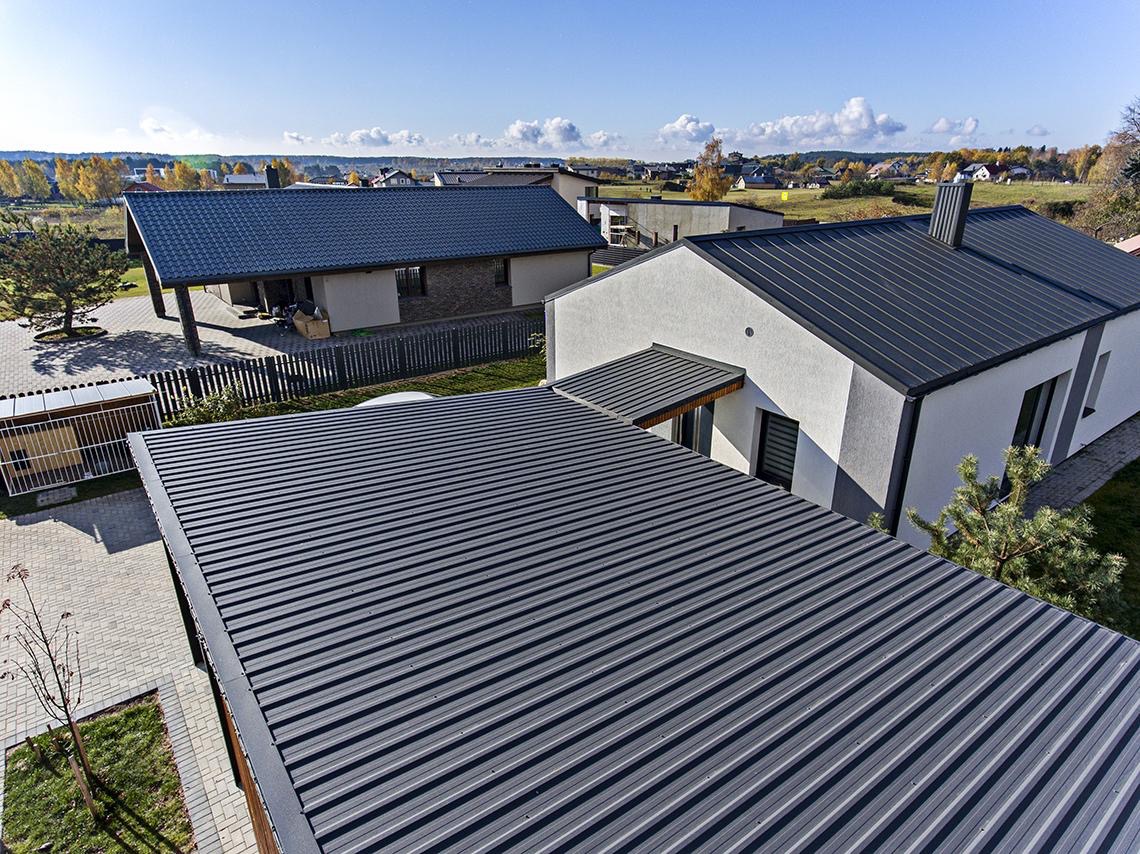 Widok z góry na nowo pokryty, ciemnoszary blaszany dach z wyraźnymi przetłoczeniami, na tle nowoczesnego domu z białymi ścianami i szarymi akcentami, w oddali inne budynki mieszkalne i jesienne...