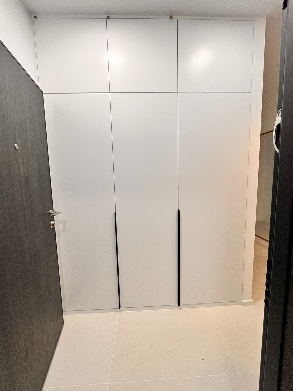 Biała szafa wnękowa z czarnymi, pionowymi uchwytami, widoczna obok ciemnoszarych drzwi wejściowych, na jasnej podłodze z płytek.