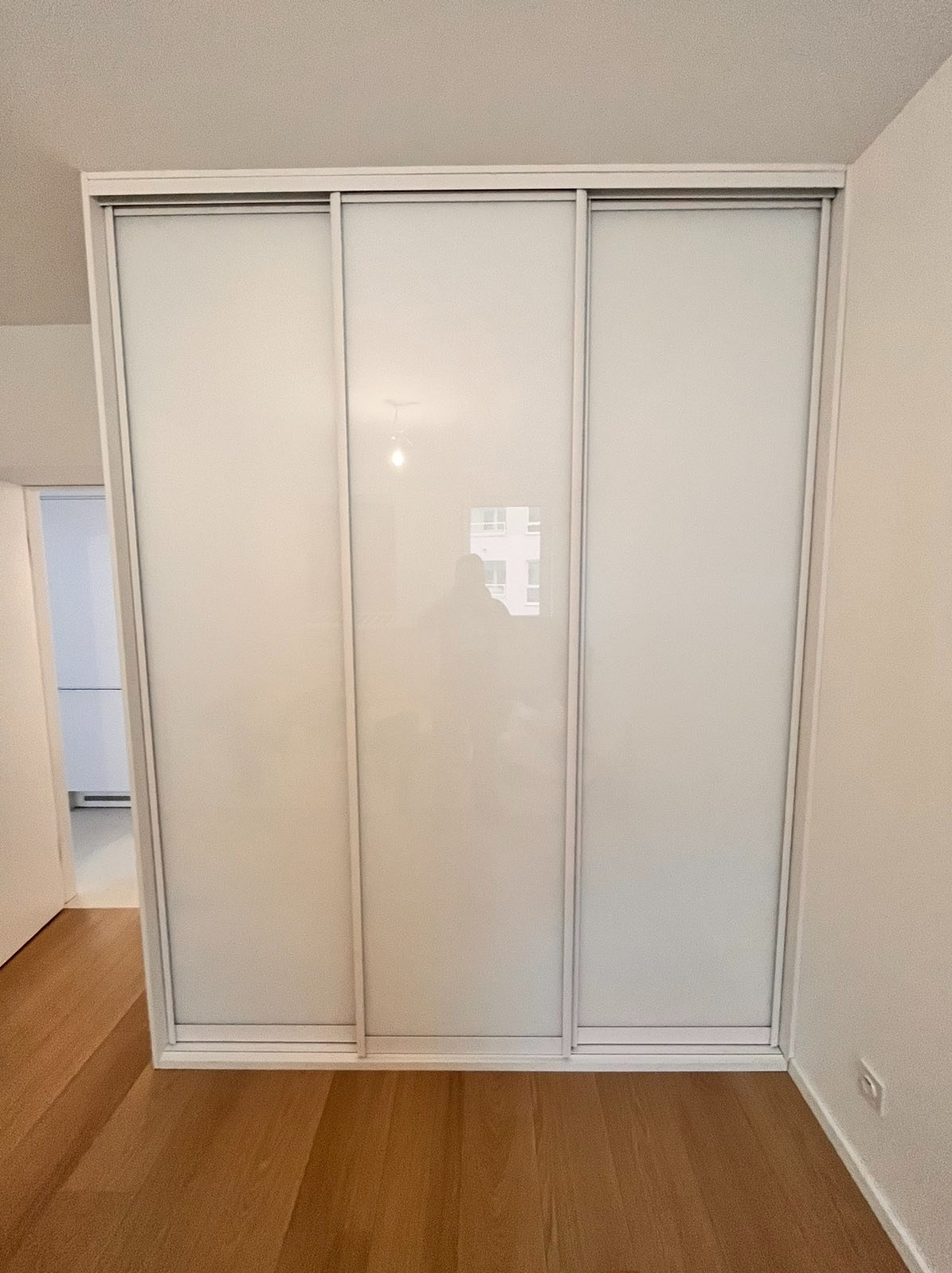 Wbudowana szafa z trzema przesuwnymi, mlecznymi drzwiami w minimalistycznym wnętrzu z parkietem. Widoczne delikatne odbicie sylwetki fotografa na środkowych drzwiach.