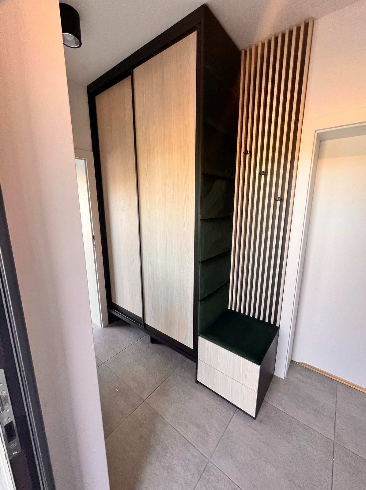 Szafa przesuwna z jasnymi frontami i czarną ramą, połączona z siedziskiem z zieloną tapicerką i panelem ściennym z drewnianych listew z wieszakami, zrealizowana w mieszkaniu w Warszawie.