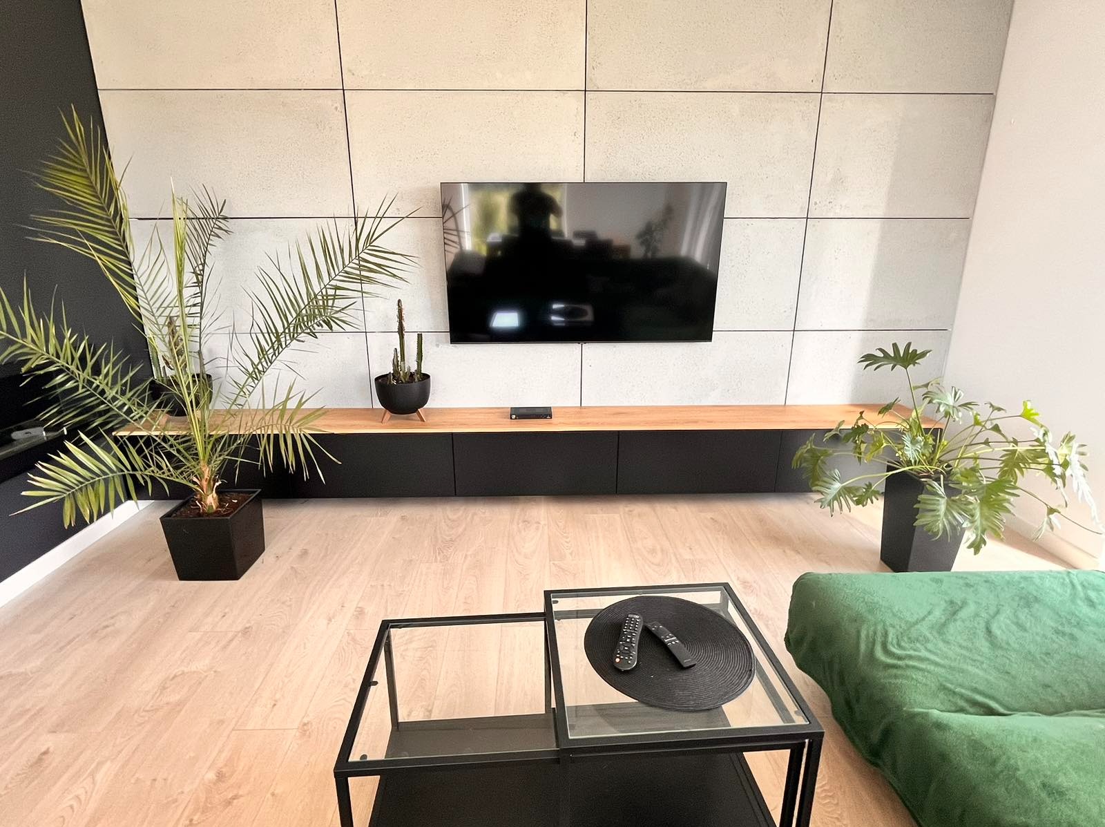 Salon z zamontowaną pod telewizorem szafką RTV z drewnianym blatem, czarnymi frontami i betonową ścianą w tle, uzupełniony roślinami doniczkowymi i czarnym stolikiem kawowym.