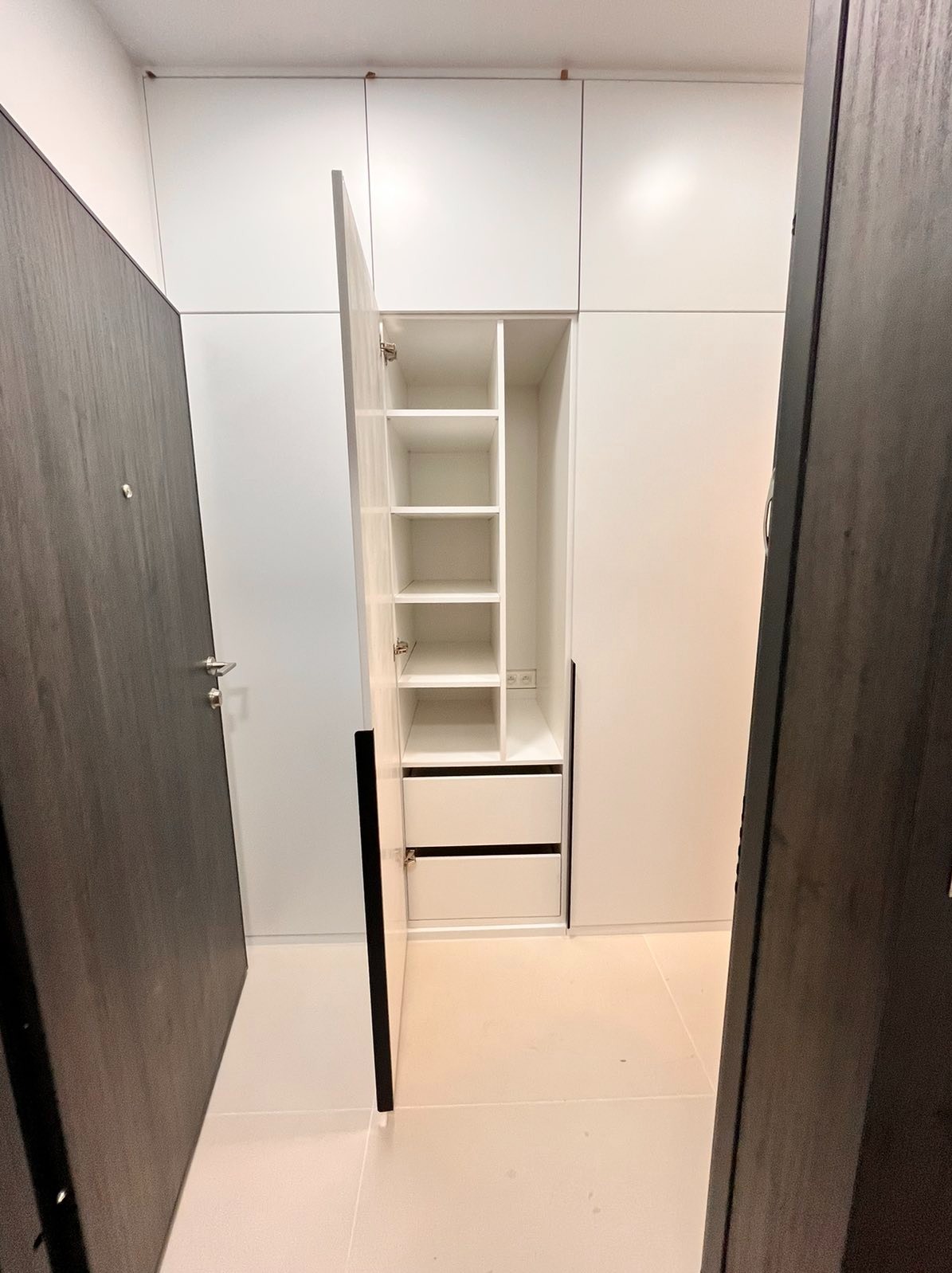 Wnękowa zabudowa meblowa w przedpokoju z otwartą szafą z półkami i szufladami, widoczna obok ciemna stolarka drzwiowa.