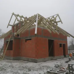 Surowy stan domu jednorodzinnego w trakcie budowy z czerwonej cegły, z częściowo zamontowaną drewnianą konstrukcją dachu, na zaśnieżonym terenie w pochmurny dzień.