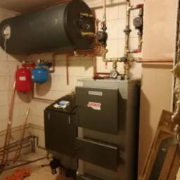 Kotłownia z kotłem na paliwo stałe marki Jarkot, zasobnikiem ciepłej wody, instalacją z miedzianych rur, manometrami i zaworami, oraz dwoma zbiornikami wyrównawczymi w kolorach czerwonym i niebieskim.