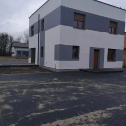 Dwukondygnacyjny dom o prostej bryle z elewacją w pasy szarości i bieli, brązowe okna z roletami, kostka brukowa na podjeździe częściowo zasypana piaskiem, drugi budynek w tle.