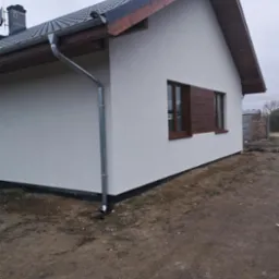 Narożne ujęcie parterowego domu z białą elewacją, ciemną dachówką, drewnianą podbitką, brązowymi oknami i metalową rynną spustową.