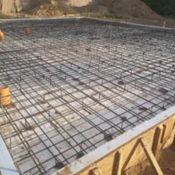 Zbrojenie płyty fundamentowej z widocznym szalunkiem, izolacją termiczną i wystającymi rurami wentylacyjnymi, na tle pryzmy ziemi.