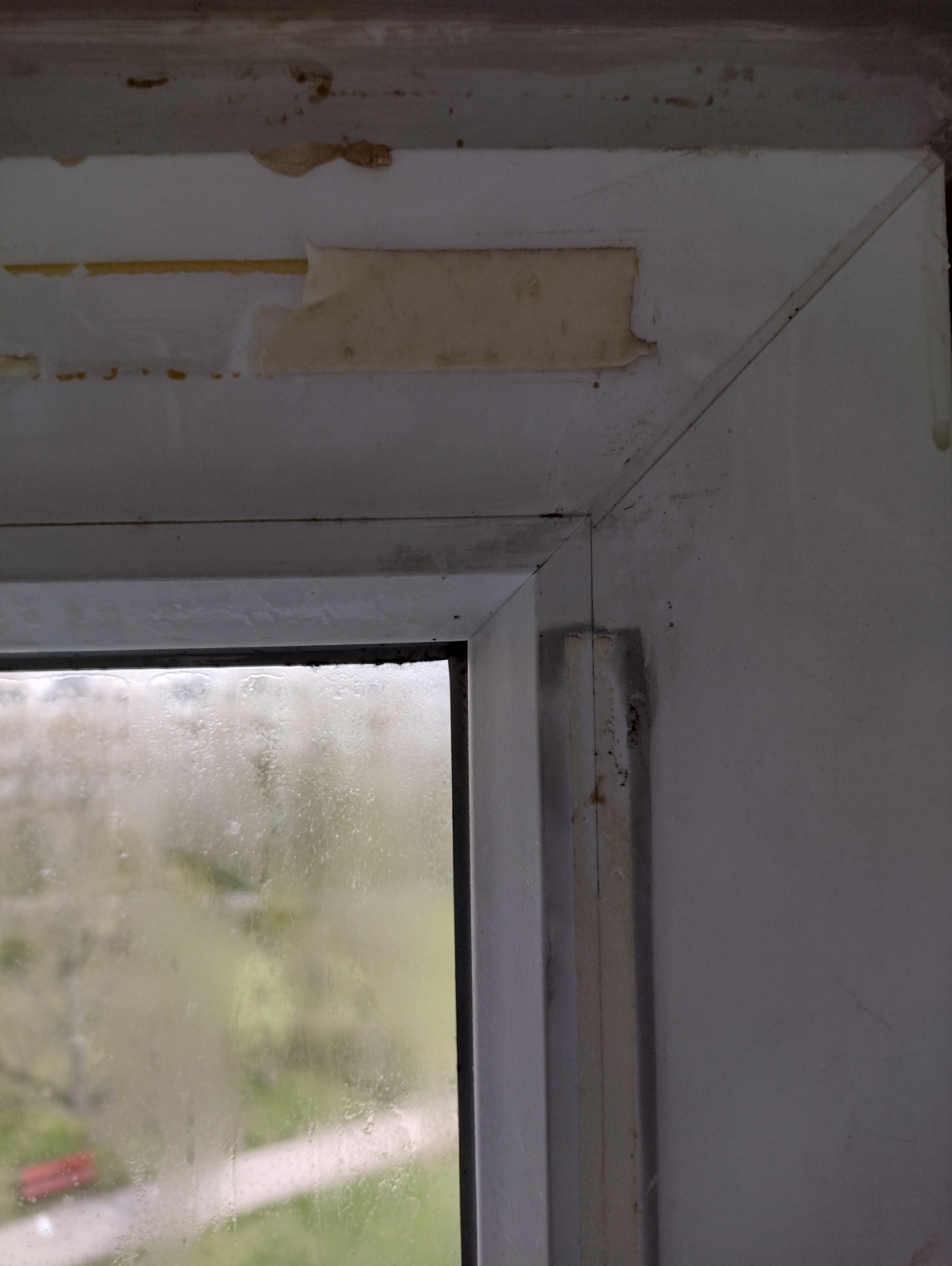 Fragment okna z widocznymi zabrudzeniami, resztkami taśmy malarskiej i zaciekami na szybie, ukazujący stan 'przed' czyszczeniem lub remontem.