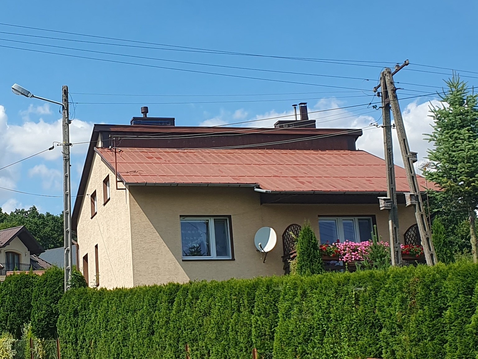 Dom jednorodzinny z czerwonym dachem widziany zza żywopłotu, z widoczną anteną satelitarną i skrzynkami z kwiatami na balkonie.