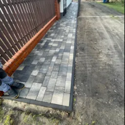 Ułożona, szara kostka brukowa prowadząca wzdłuż brązowego płotu z ceglaną podmurówką, widoczne buty pracownika w lewym dolnym rogu.