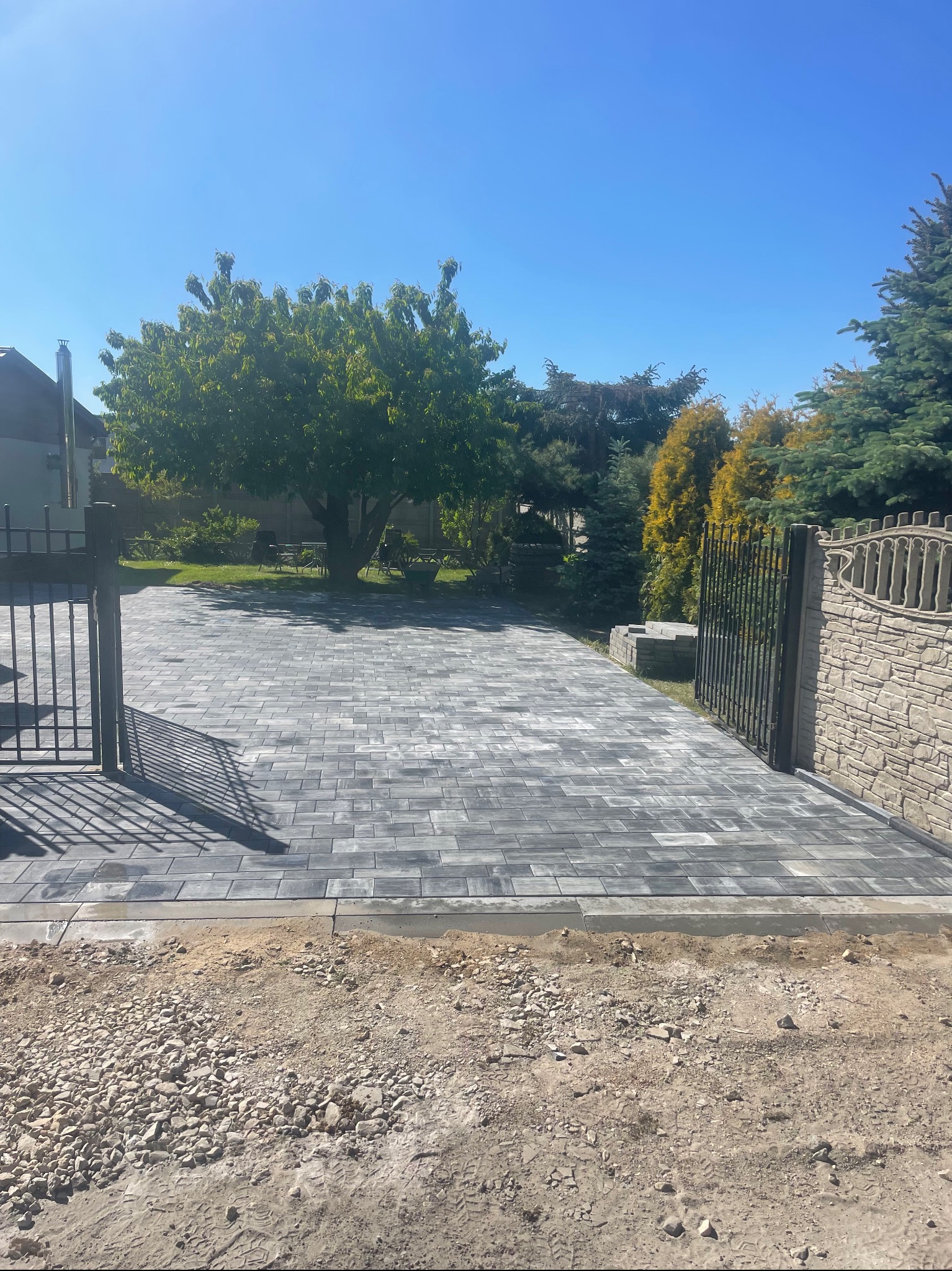 Ułożona szara kostka brukowa na podjeździe prowadzącym do domu, widoczny fragment ogrodzenia i brama, z pozostawionym pasem ziemi z kamieniami do wykończenia.