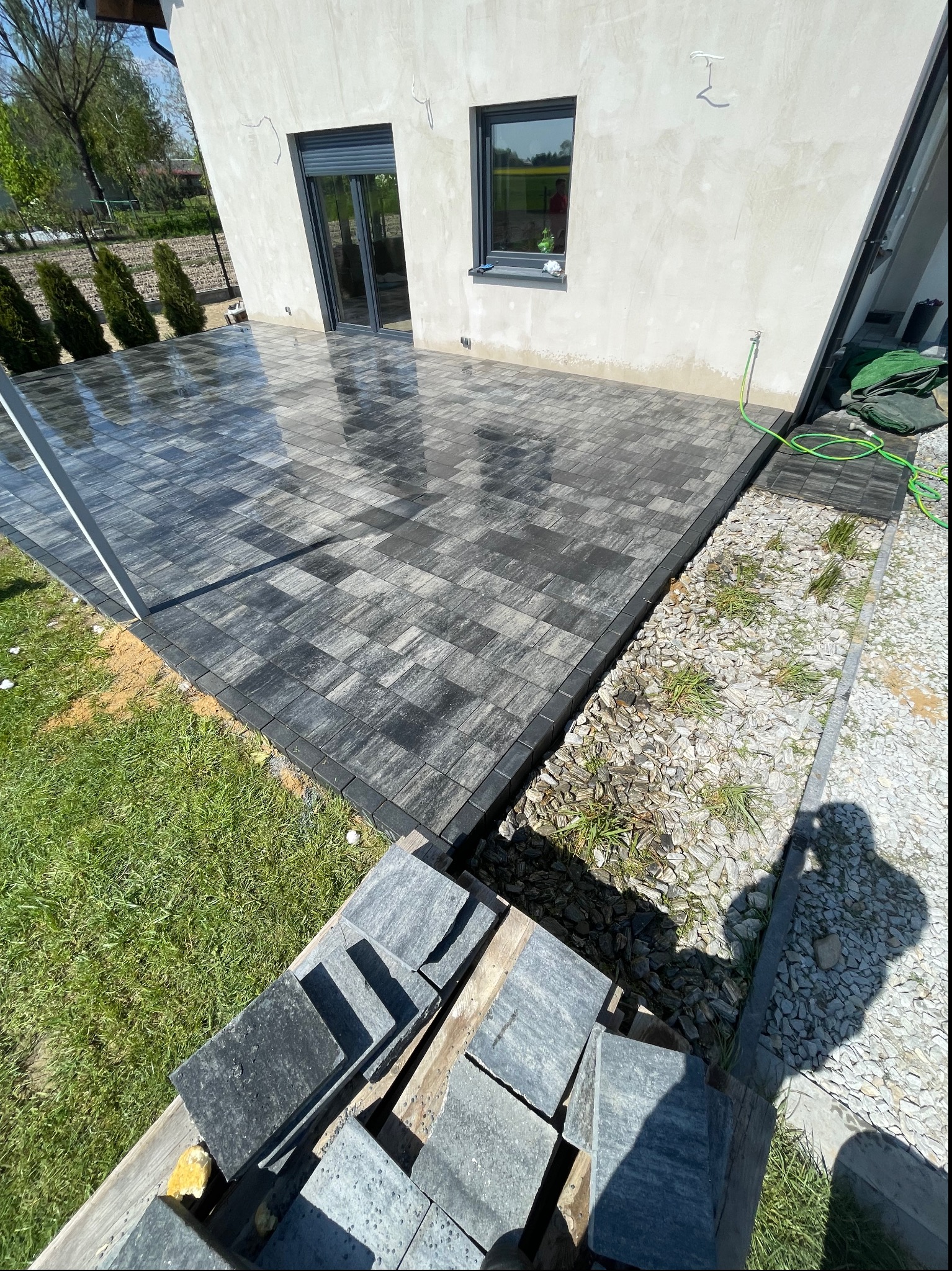 Świeżo ułożona, mokra kostka brukowa w odcieniach szarości, przylegająca do jasnego budynku z szarymi oknami; obok rabata z kamykami i zielenią oraz paleta z zapasem kostki.