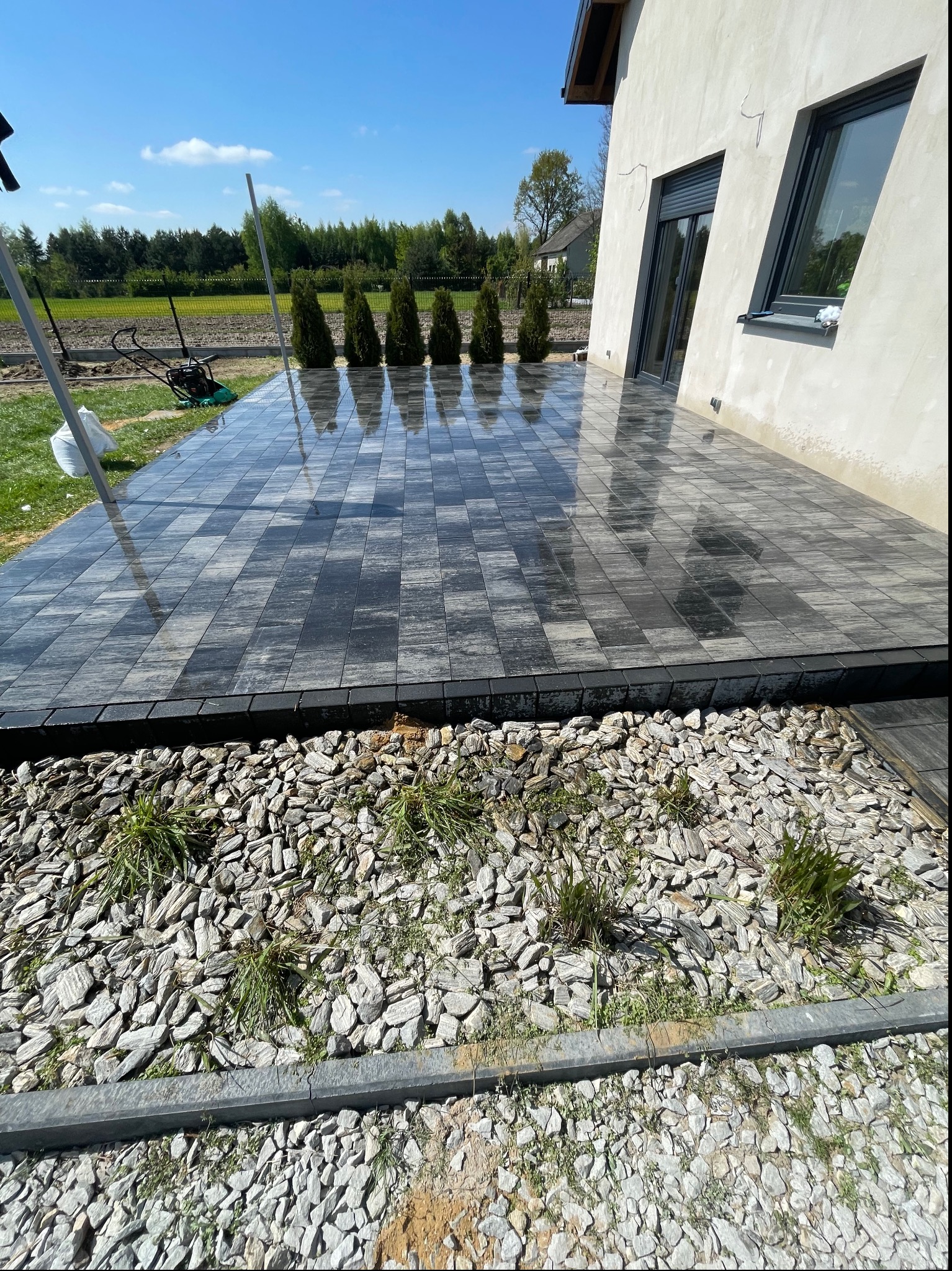 Nowo ułożony taras z szarej kostki brukowej o regularnym wzorze, sąsiadujący z rabatą wysypaną białym gresem. W tle widoczne tuje i trawnik, a na nim sprzęt budowlany.