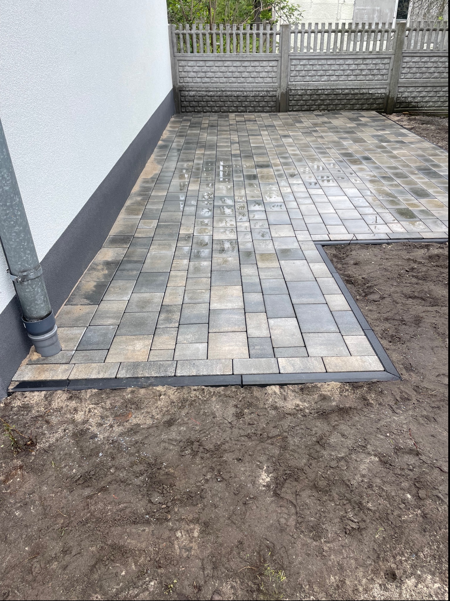 Świeżo ułożona, mokra kostka brukowa w odcieniach szarości, przylegająca do ściany budynku z rynną spustową i betonowego ogrodzenia. Fragment terenu bez kostki, obramowany czarnym obrzeżem.
