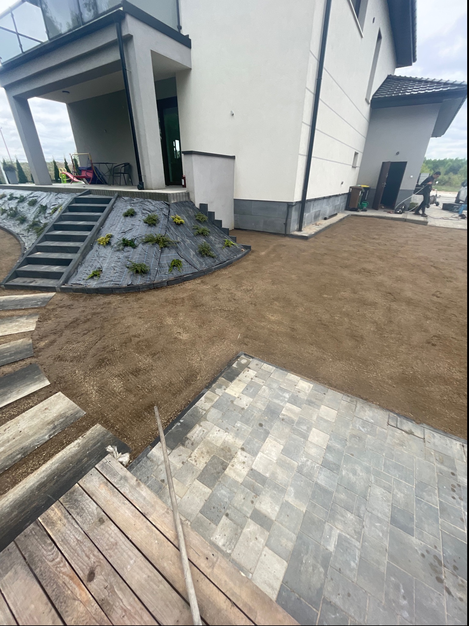 Teren wokół domu w trakcie prac ziemnych: ułożona częściowo kostka brukowa, drewniane schody, nasyp z roślinami, przygotowany teren pod trawnik.