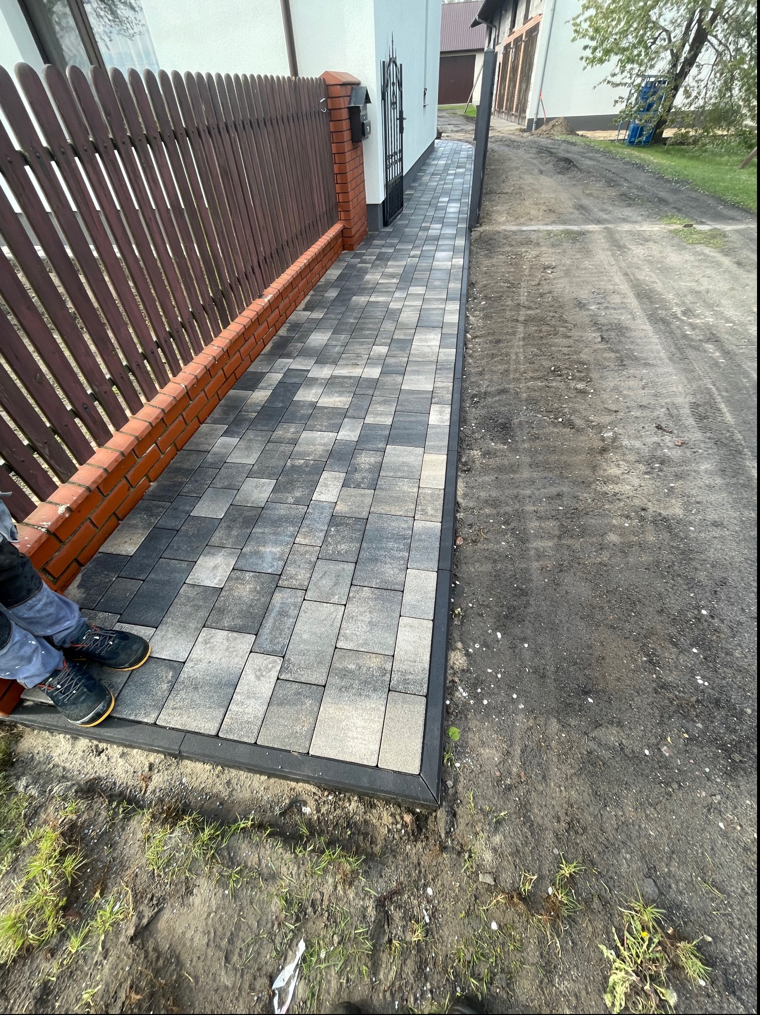 Ułożona, szara kostka brukowa prowadząca wzdłuż brązowego płotu z ceglaną podmurówką, widoczne buty pracownika w lewym dolnym rogu.