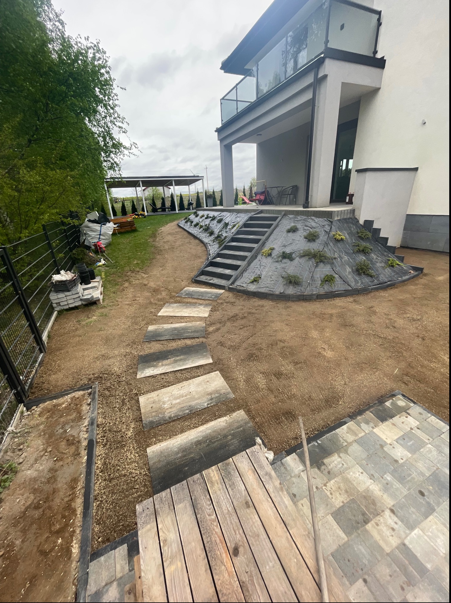 Nowoczesny dom z ogrodem w trakcie aranżacji: ułożona ścieżka z płyt imitujących drewno, schody z ciemnej kostki prowadzące do domu, częściowo zagospodarowany teren z roślinami na skarpie, widoczne...