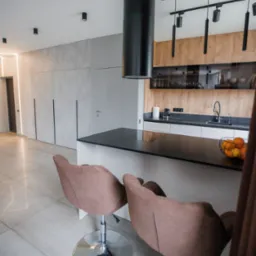 Nowoczesna kuchnia z wyspą i hokerami, czarny okrągły okap nad wyspą, oświetlenie szynowe i szafki z drewnianymi frontami w tle.