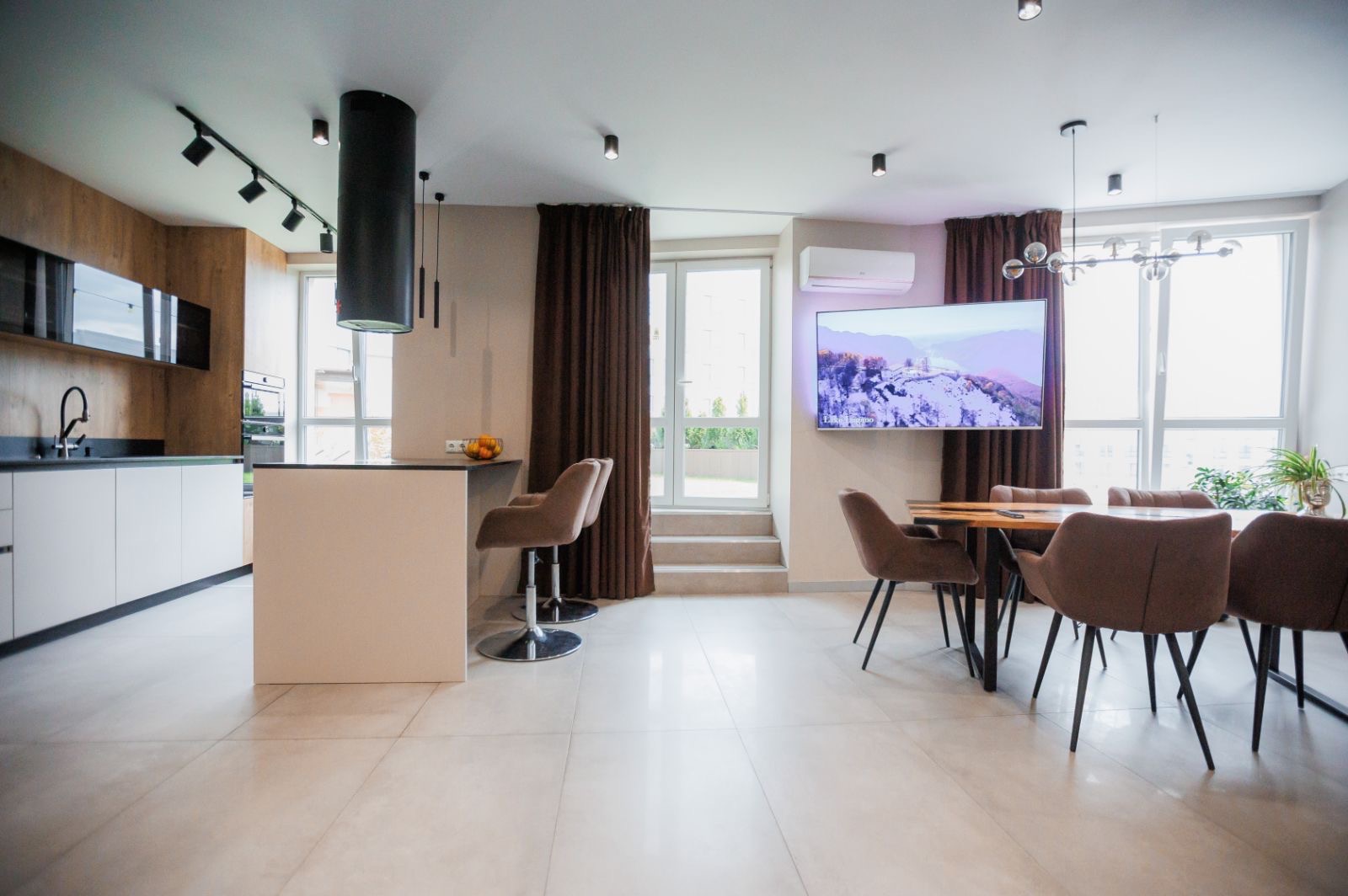 Nowoczesne wnętrze: otwarta kuchnia z wyspą, hokery, stół jadalny z krzesłami, telewizor na ścianie, jasne płytki podłogowe i designerskie oświetlenie.