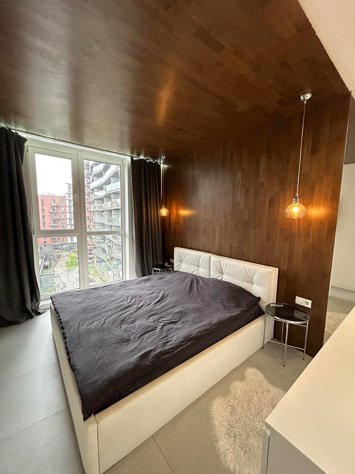 Sypialnia z drewnianą ścianą, białym łóżkiem i szarą pościelą, oświetlona wiszącymi lampami z żarówkami Edisona, widok na osiedle przez duże okno.