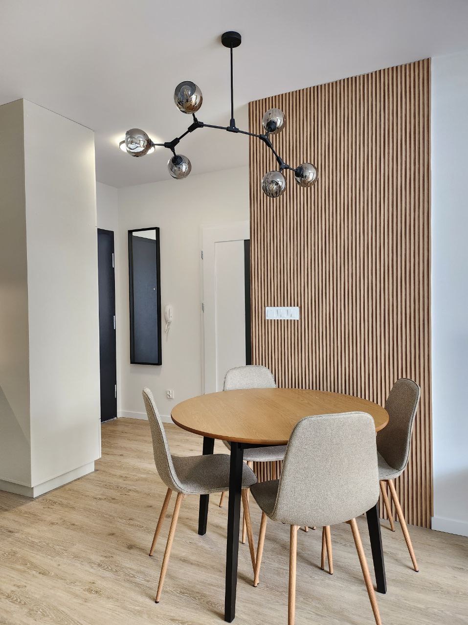 Jasne wnętrze jadalni z okrągłym drewnianym stołem na czarnej nodze, czterema szarymi krzesłami, drewnianą ścianą z pionowych listew i nowoczesną lampą sufitową z sześcioma szklanymi kloszami.