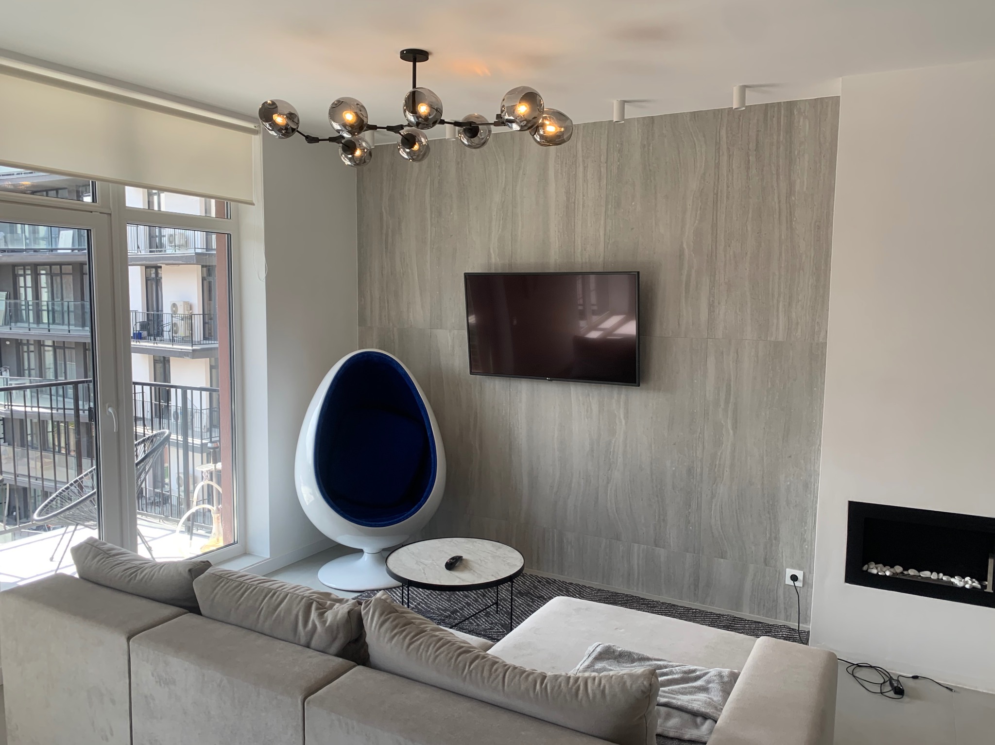 Nowoczesny salon z szarą, teksturowaną ścianą za telewizorem, designerski fotel w kształcie jaja z granatowym wnętrzem, geometryczny dywan, minimalistyczny kominek i duży, modułowy narożnik...