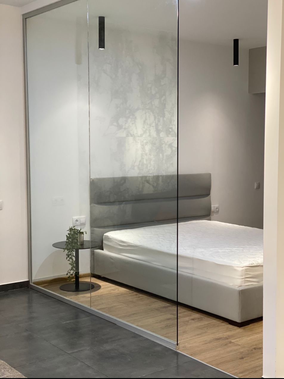 Nowoczesna sypialnia oddzielona szklaną ścianą, widoczny fragment łóżka z szarym zagłówkiem i białą pościelą, mały stolik nocny z rośliną, minimalistyczne czarne lampy sufitowe.