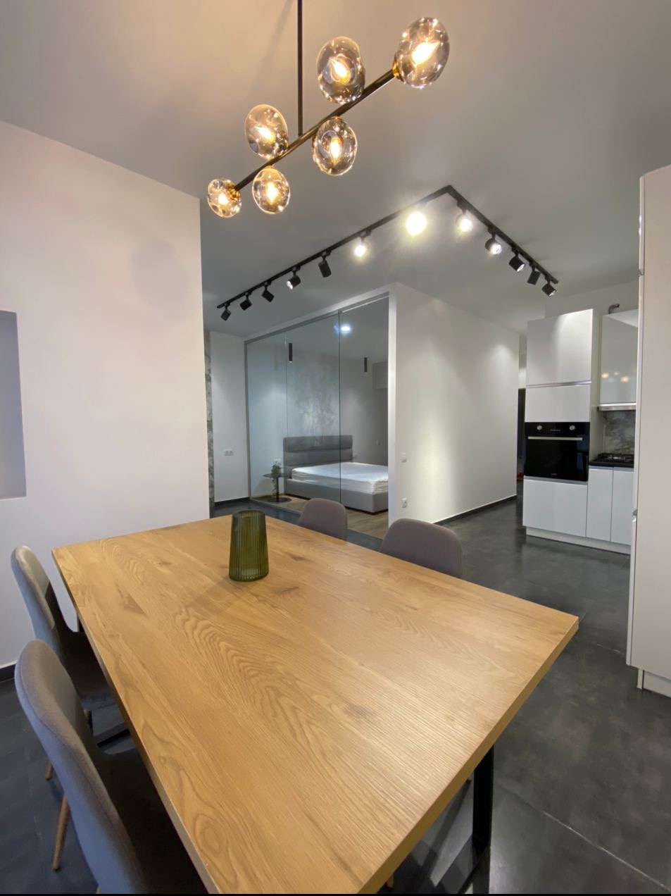 Nowoczesne wnętrze: drewniany stół jadalny z szarymi krzesłami, szklana ściana oddzielająca sypialnię z szarym łóżkiem, oświetlenie sufitowe punktowe i designerska lampa wisząca, biała zabudowa...