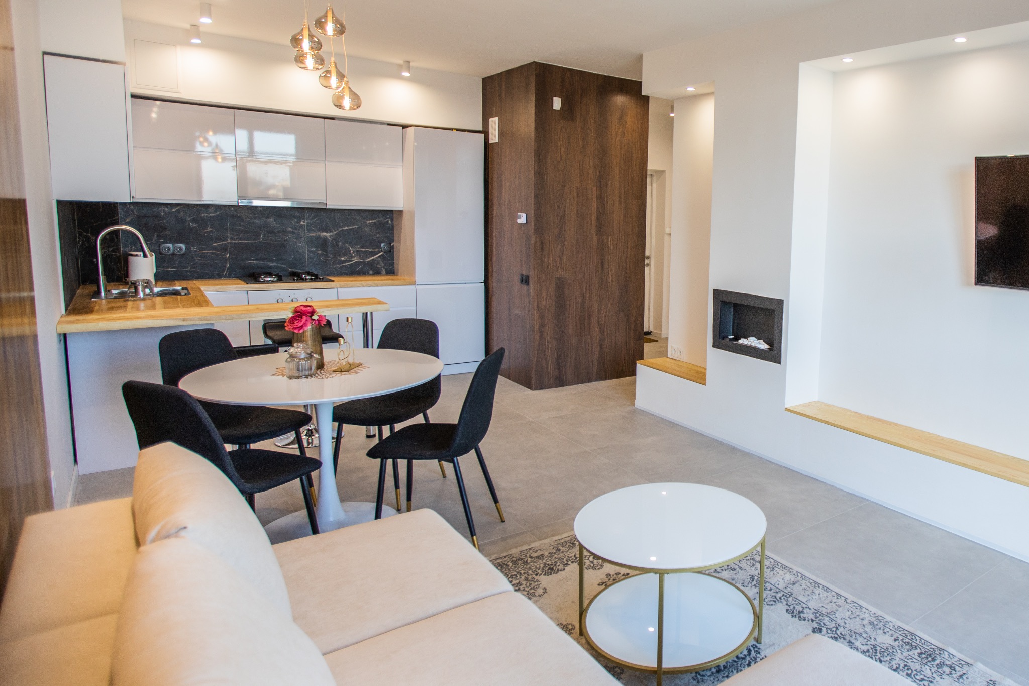 Nowoczesne wnętrze: kuchnia z czarnym marmurem nad blatem, okrągły stół jadalny z czarnymi krzesłami, minimalistyczny kominek w ścianie i beżowa sofa w salonie.