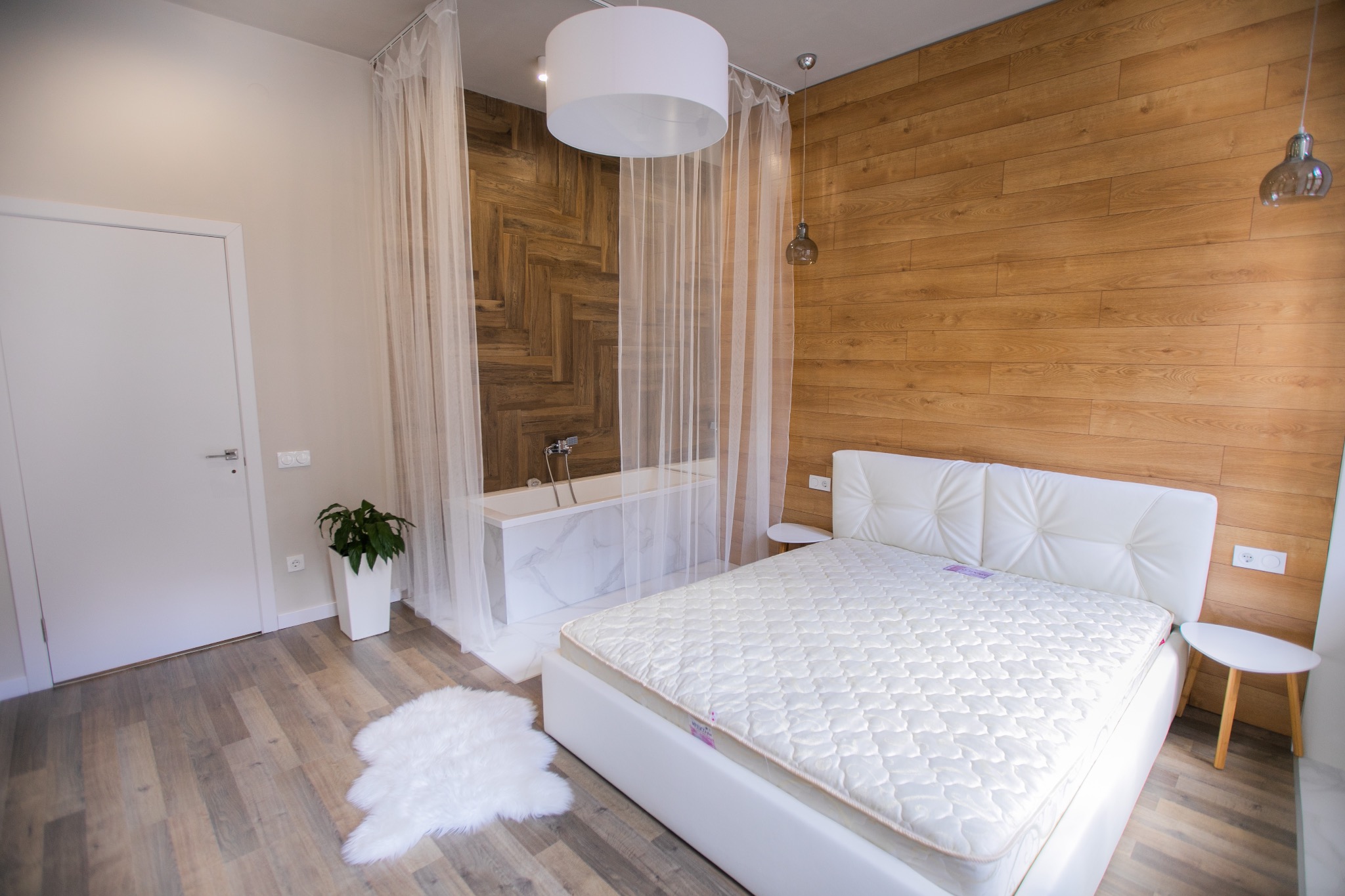 Jasna sypialnia z drewnianą ścianą, białym łóżkiem i wanną za zwiewną zasłoną, oświetlona nowoczesną lampą.