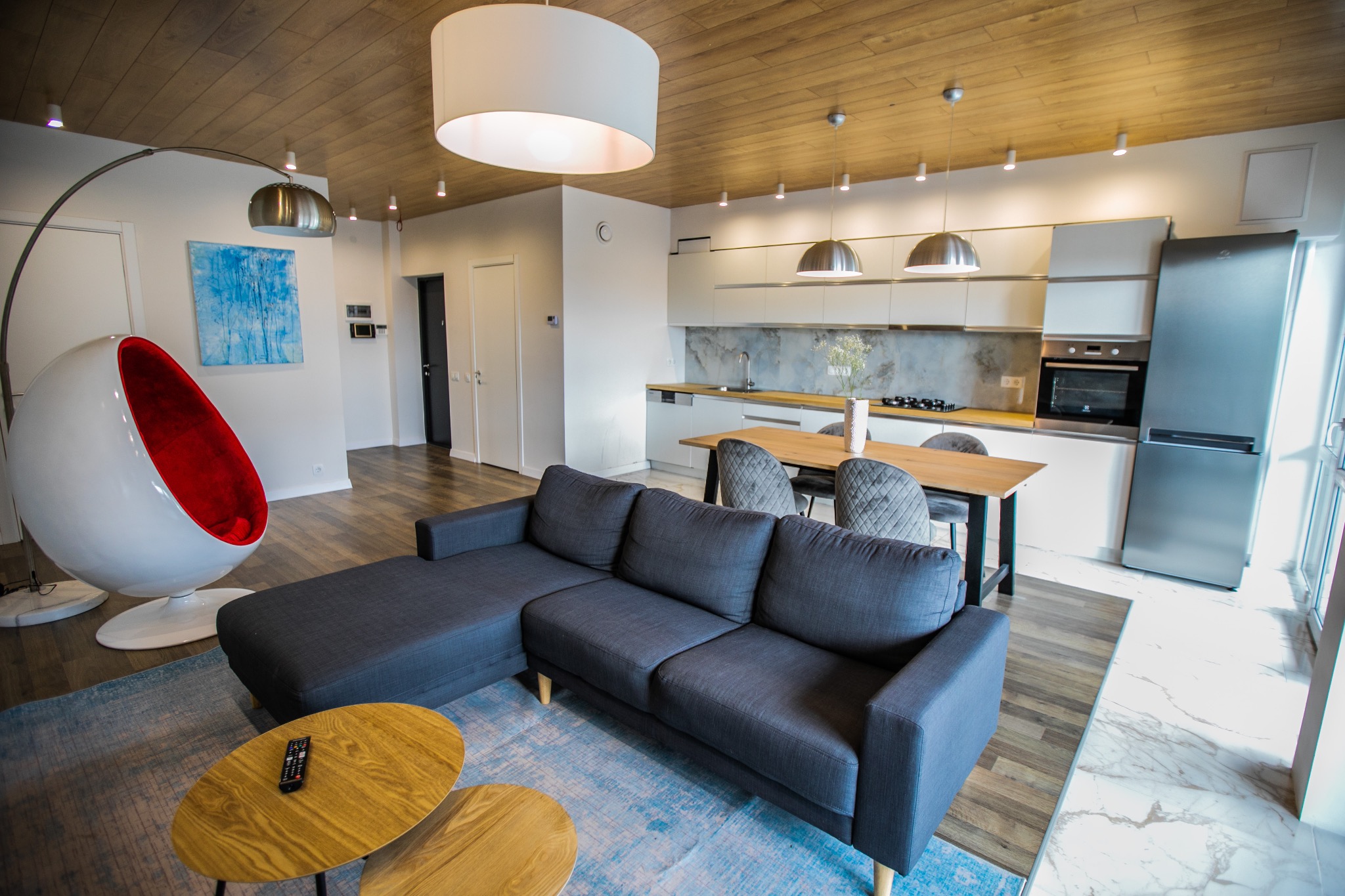 Nowoczesne wnętrze mieszkania z szarą sofą narożną, drewnianym stołem i białymi szafkami kuchennymi pod drewnianym sufitem.