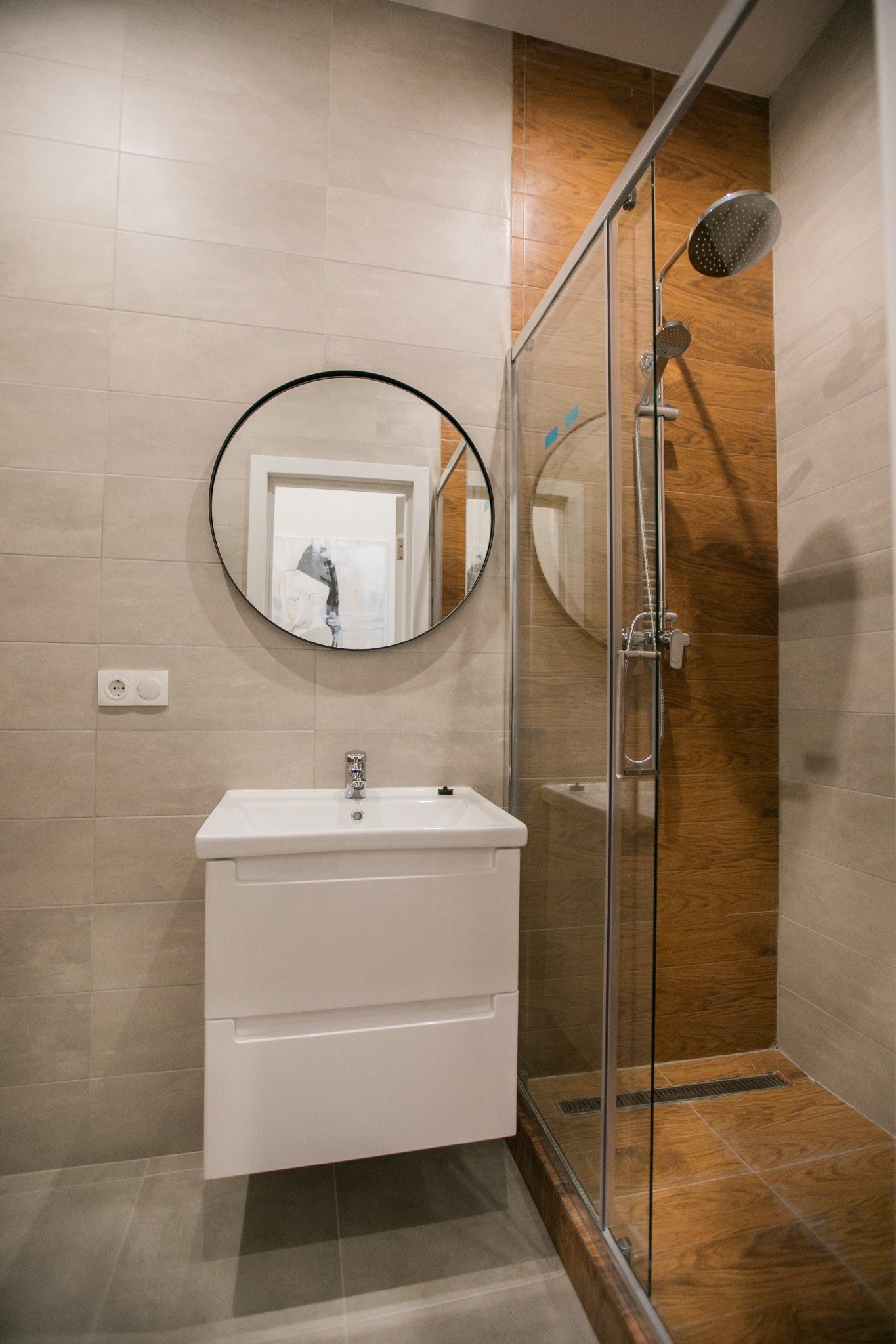Nowoczesna łazienka z kabiną prysznicową typu walk-in, umywalką z szafką i okrągłym lustrem; widoczne płytki imitujące drewno w strefie prysznica i beżowe płytki na pozostałych ścianach.