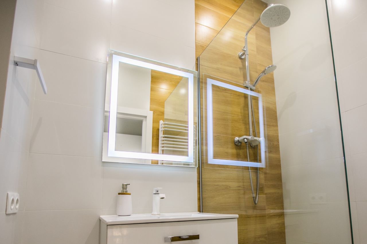 Nowoczesna łazienka z białymi płytkami, drewnopodobną strefą prysznicową, podświetlanym lustrem i białą umywalką z dozownikiem mydła.