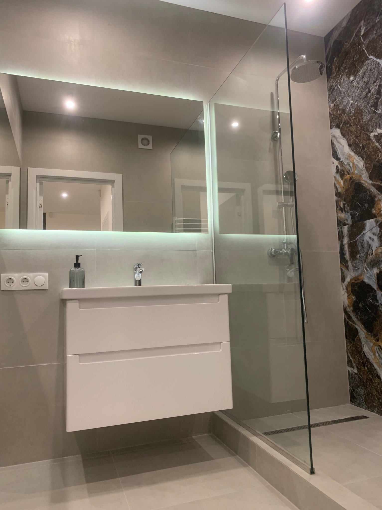 Nowoczesna łazienka z białą szafką umywalkową, podświetlanym lustrem, szklaną kabiną prysznicową i ścianą z płytek imitujących marmur.