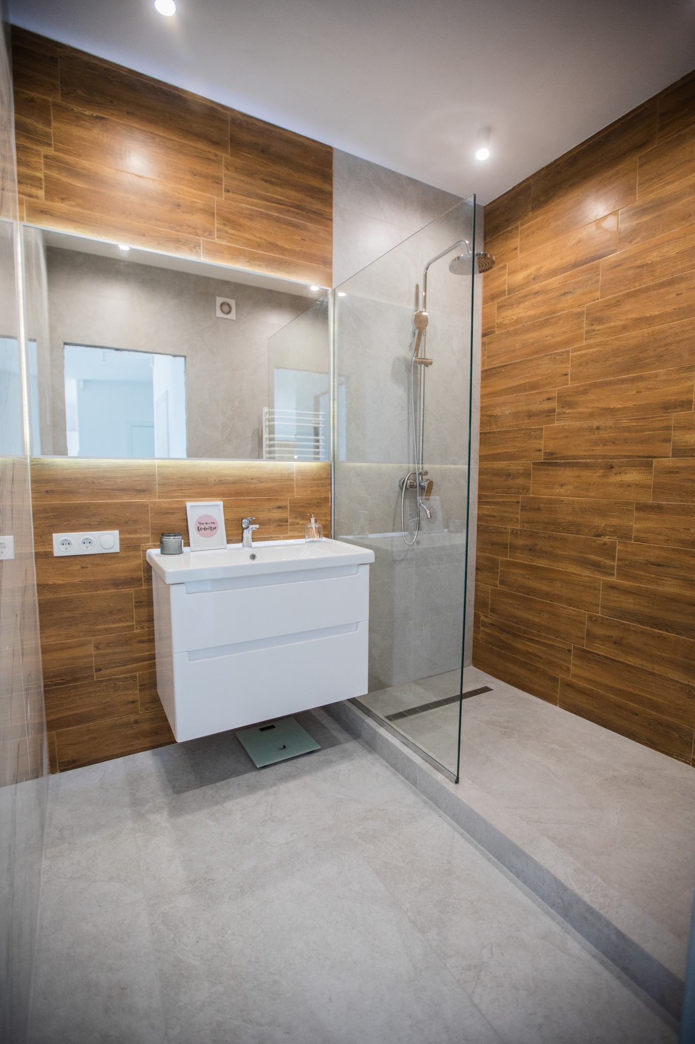 Nowoczesna łazienka z drewnopodobnymi i szarymi płytkami, kabina prysznicowa typu walk-in, biała szafka pod umywalką z lustrem z podświetleniem LED i dekoracjami na blacie, w tle widoczna waga...