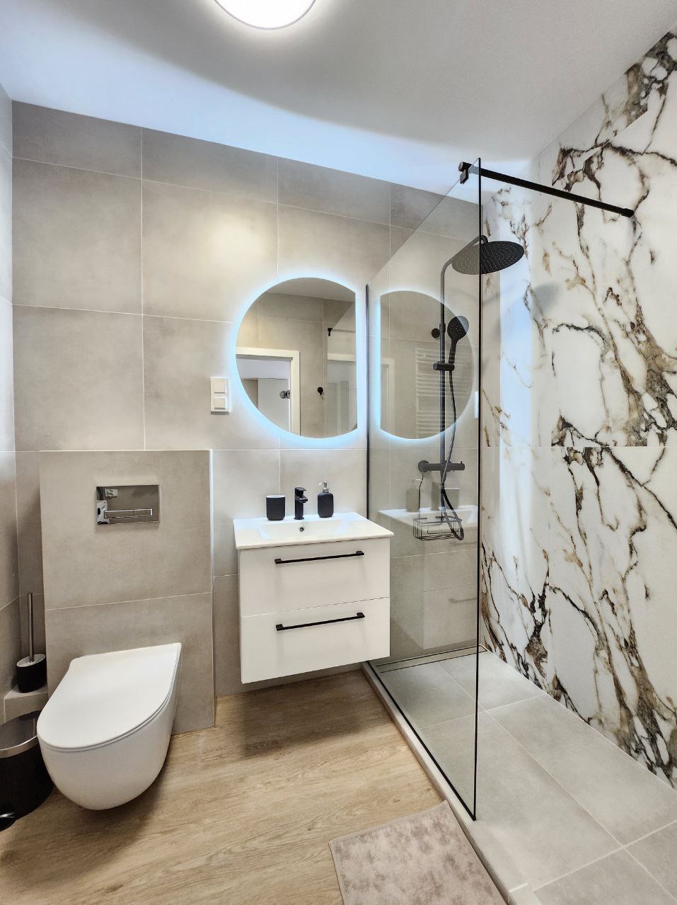 Nowoczesna łazienka z kabiną prysznicową typu walk-in, okrągłym lustrem z podświetleniem LED, białą szafką pod umywalką z czarnymi uchwytami i imitacją marmuru na ścianie.