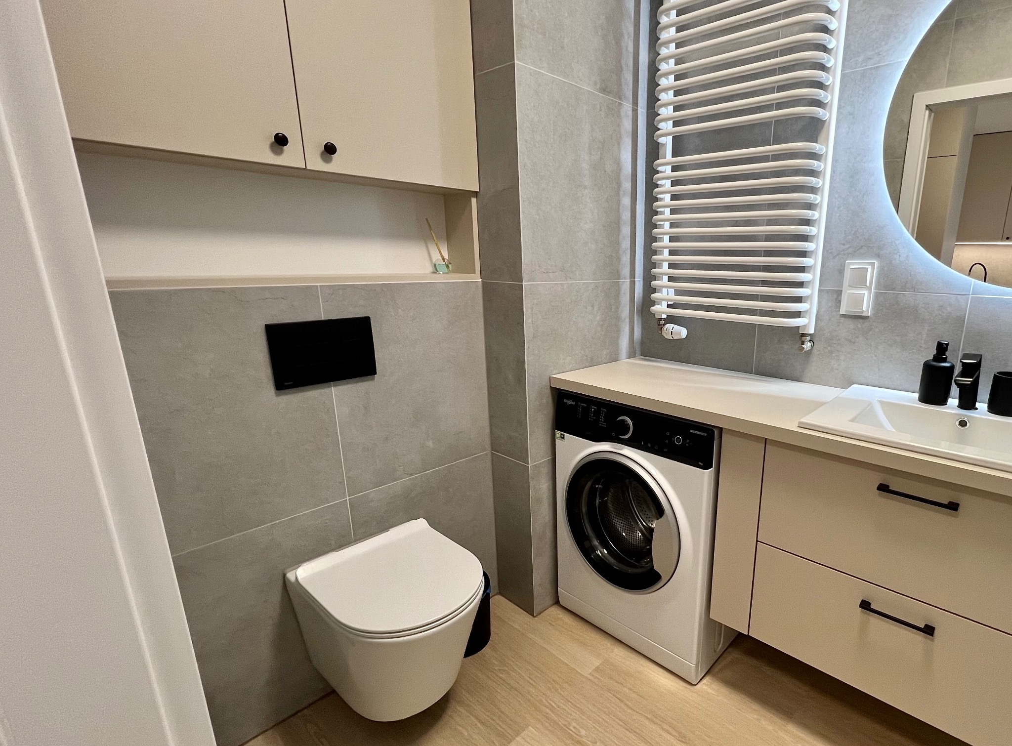 Jasna łazienka z szarymi płytkami, wiszącą toaletą, pralką pod blatem umywalki i okrągłym lustrem z podświetleniem LED.