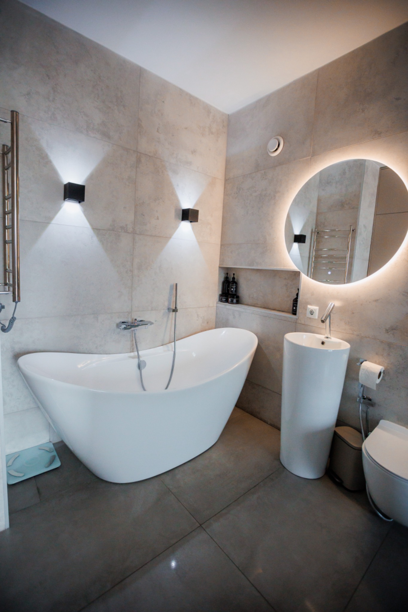 Nowoczesna łazienka w Krakowie z wolnostojącą wanną, podświetlanym lustrem i szarą płytką imitującą beton na ścianach, oświetlona czarnymi kinkietami.