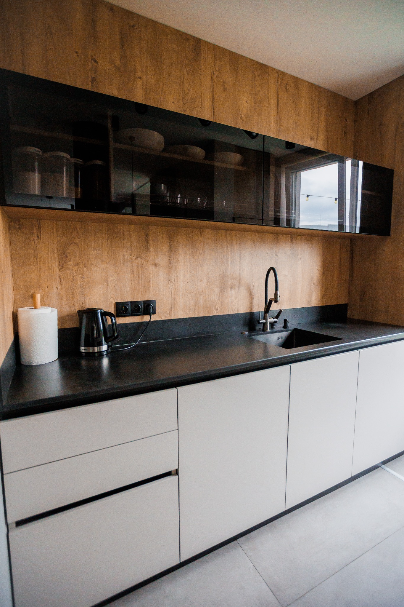 Nowoczesna kuchnia z drewnianą okładziną ściany, czarnym blatem i szarymi szafkami, wyposażona w czarny zlew, baterię, czajnik i pojemnik na ręczniki papierowe.