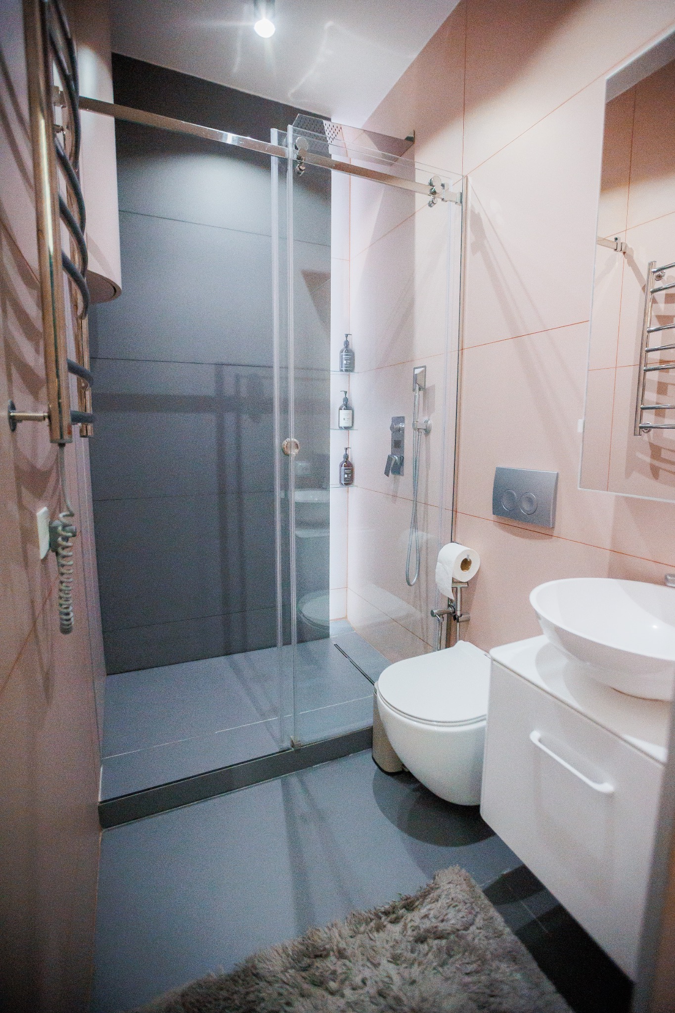 Nowoczesna łazienka z kabiną prysznicową ze szklanymi drzwiami przesuwnymi, grzejnikiem drabinkowym i umywalką nablatową na białej szafce, wykończona płytkami w odcieniach różu i szarości,...